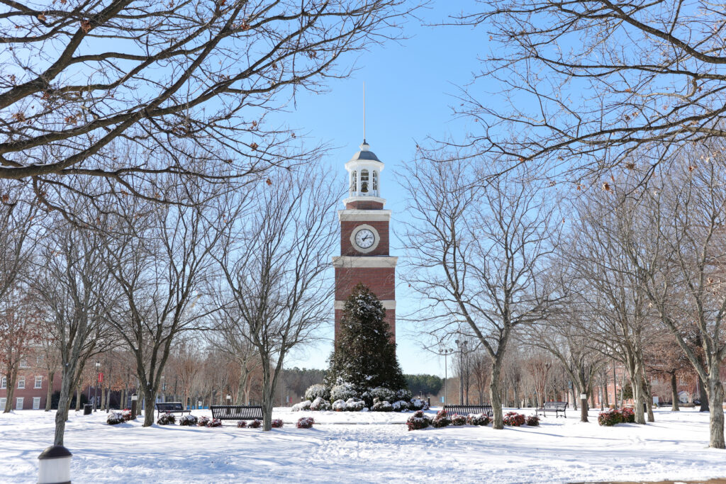 A brick clock tower stands at the center of Tennessee’s snow-covered Union main campus in Jackson, surrounded by leafless trees, benches, and a large evergreen under a clear blue sky.
