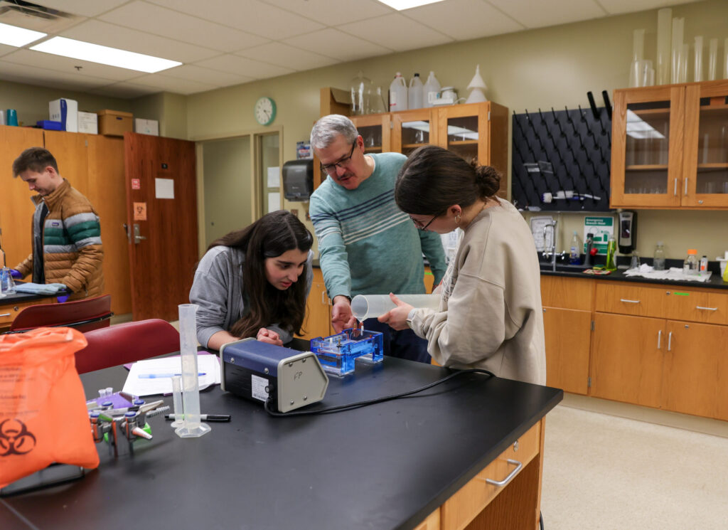 Three people work together at a lab bench with scientific equipment and notes. One person instructs the others, who are focused on handling lab materials. Shelves with glassware and cabinets are visible in the background.