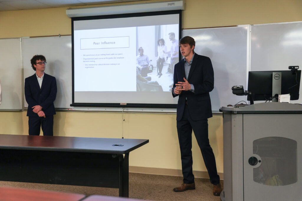 Two men in suits give a presentation on Peer Influence in a classroom. The discussion focuses on topics relevant to Management and Human Resources, with one presenter near the projector screen and a computer podium visible to the right.