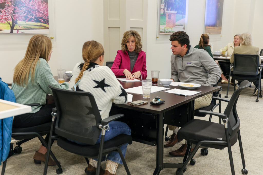 Four people sit around a black table in a meeting room at the McAfee School of Business, engaged in discussion. Papers, glasses, and plates are on the table. Two others are visible in the background at another table.