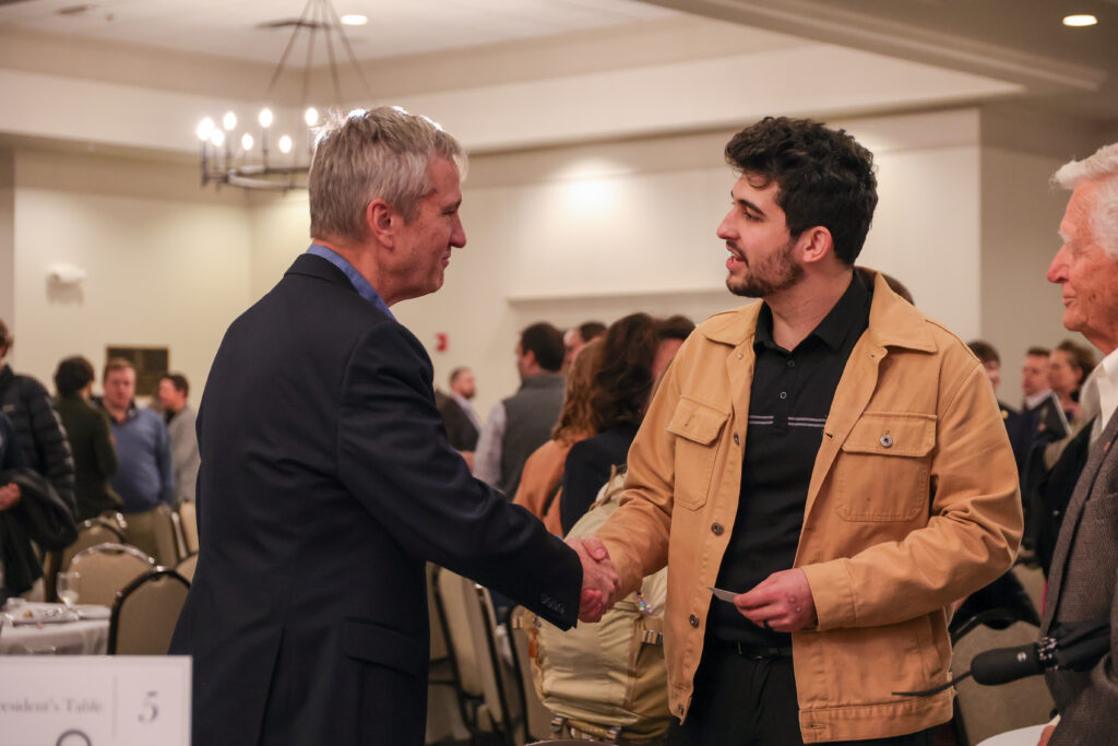 Two men are shaking hands and speaking at an indoor event with several other people present in the background. Both are wearing jackets, and there is a chandelier hanging from the ceiling.