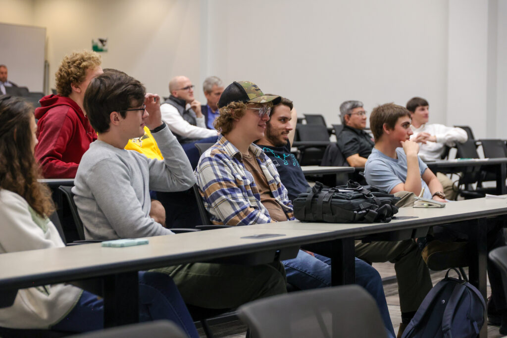 A group of people sit in rows of chairs in a classroom or lecture hall, engaged in university studies. Most are young adults, with notebooks and bags on the desks in front of them. The room has white walls and a casual atmosphere.
