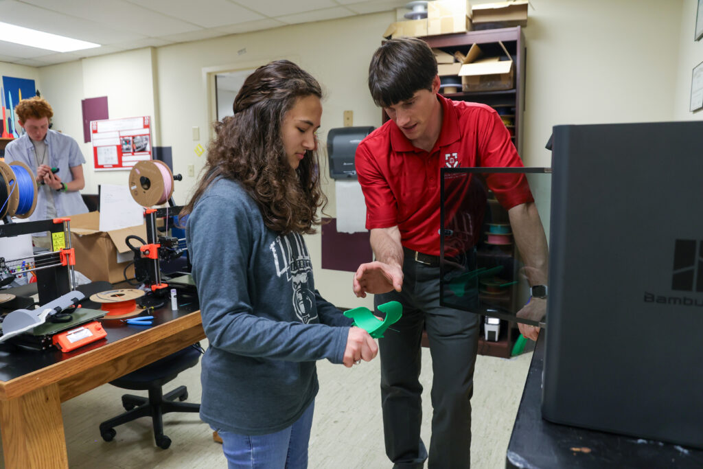 A man and a young woman stand in a room with 3D printers, discussing engineering. The man gestures toward a piece of green material the woman is holding, while another person works at a desk with spools and tools in the background.
