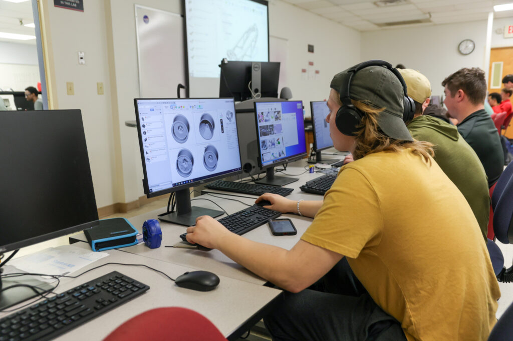 A person in a yellow shirt and headphones works on a CAD design at a computer in a classroom, engaged in Computer Engineering alongside other students also using computers.