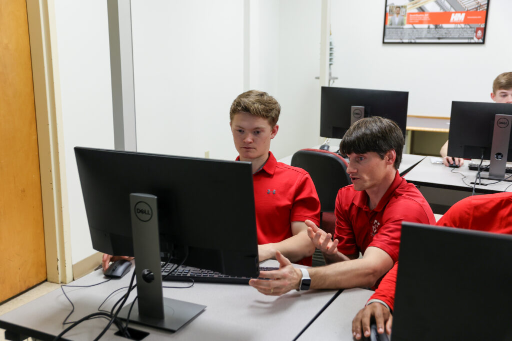 Two people sit at a computer in a classroom. One points at the screen while discussing concepts from Computational Science, while the other types on the keyboard. Both wear red shirts. Computer monitors and other students are visible in the background.