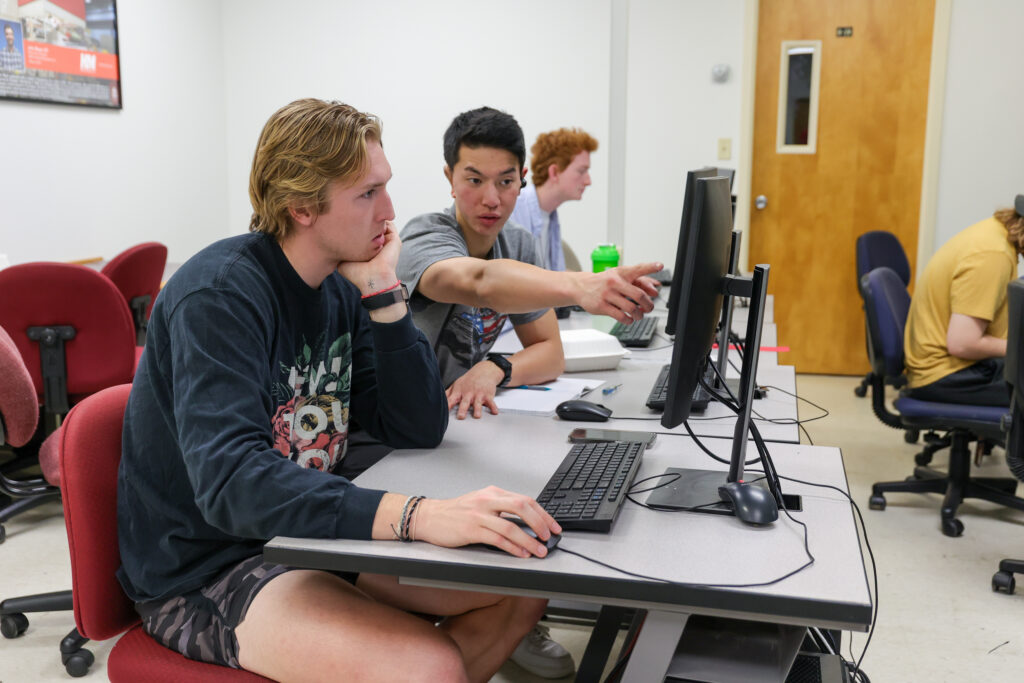 Two students sit at a desk working on a computer in a Computer Engineering classroom. One student points at the monitor while the other looks at the screen. Other students work at computers in the background.