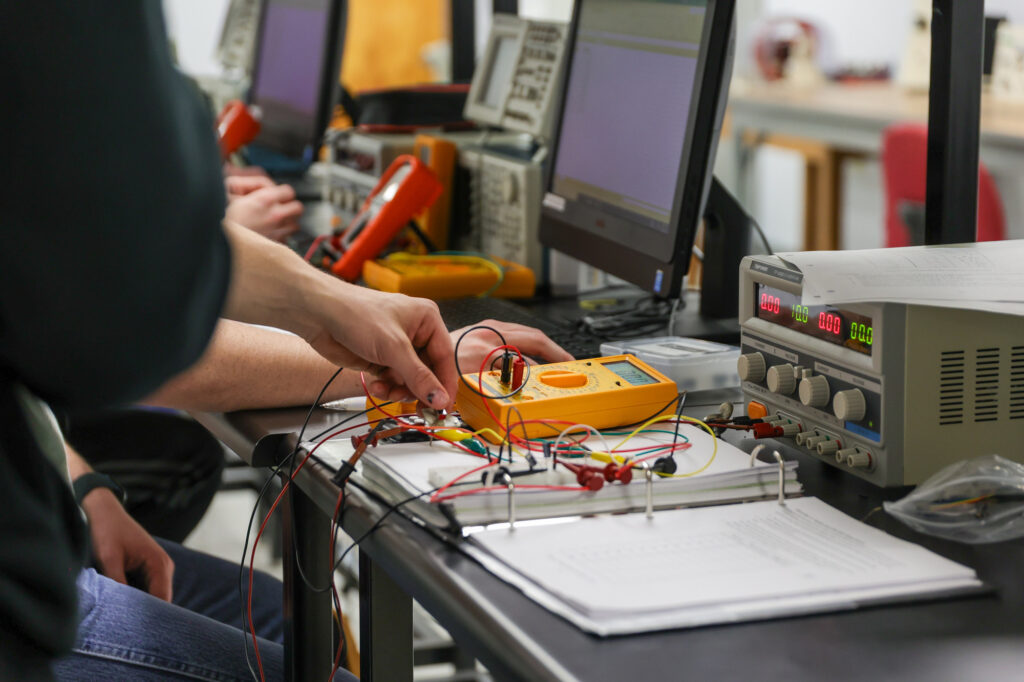 A person uses a yellow digital multimeter to test electronic components connected by wires on a lab bench, with computers and equipment visible in the background.