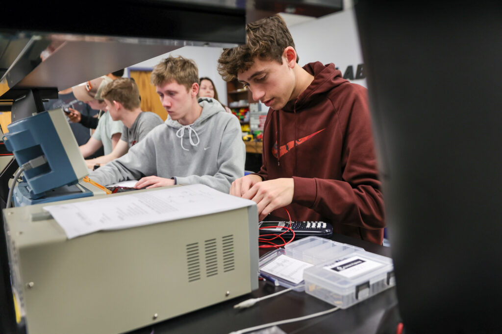 Four students work at a lab bench with electronic equipment and wiring tools. One student in a maroon hoodie focuses on connecting wires. Papers, toolboxes, and devices are spread out on the bench in front of them.