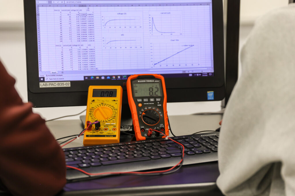 Two digital multimeters are placed on a keyboard in front of a computer monitor displaying graphs and data tables in spreadsheet software. Two people are sitting on either side of the equipment.