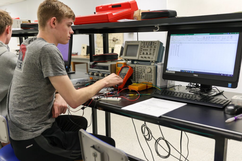 A person in a gray t-shirt uses electronic testing equipment at a lab bench, with a computer monitor displaying a spreadsheet and various instruments and wires on the desk. Another person is partially visible on the left.