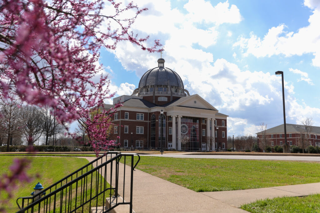 A brick building with a large dome and columns stands on Union's main campus in Jackson, Tennessee. Pink flowers bloom on a tree in the foreground, while a grassy lawn and sidewalk lead up to the building under a partly cloudy sky.