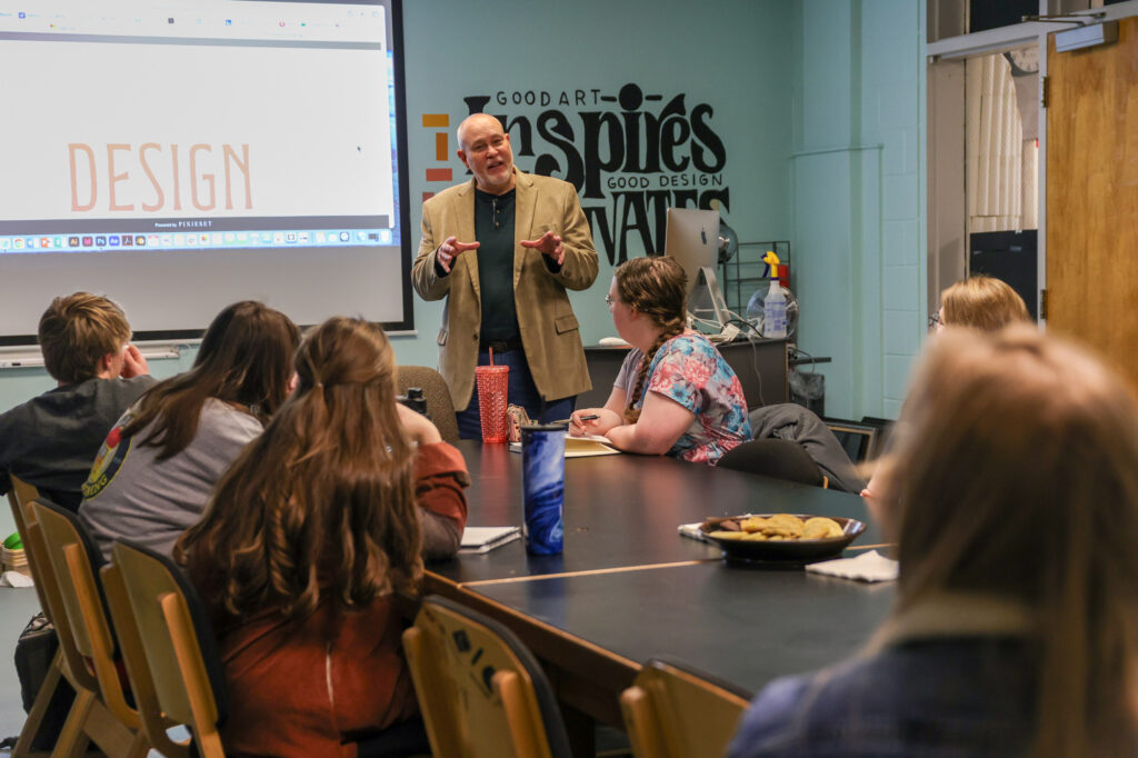 A man stands and speaks to a group of students in a classroom filled with art supplies, snacks, and a wall that reads Good art inspires. A screen behind him displays the word DESIGN, highlighting the importance of Graphic Design in their creative journey.