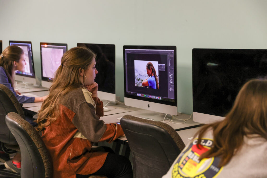Several students sit at a row of computers in a classroom. One student is editing an image of a person with photo editing software, working on a Graphic Design project. The walls are light blue.