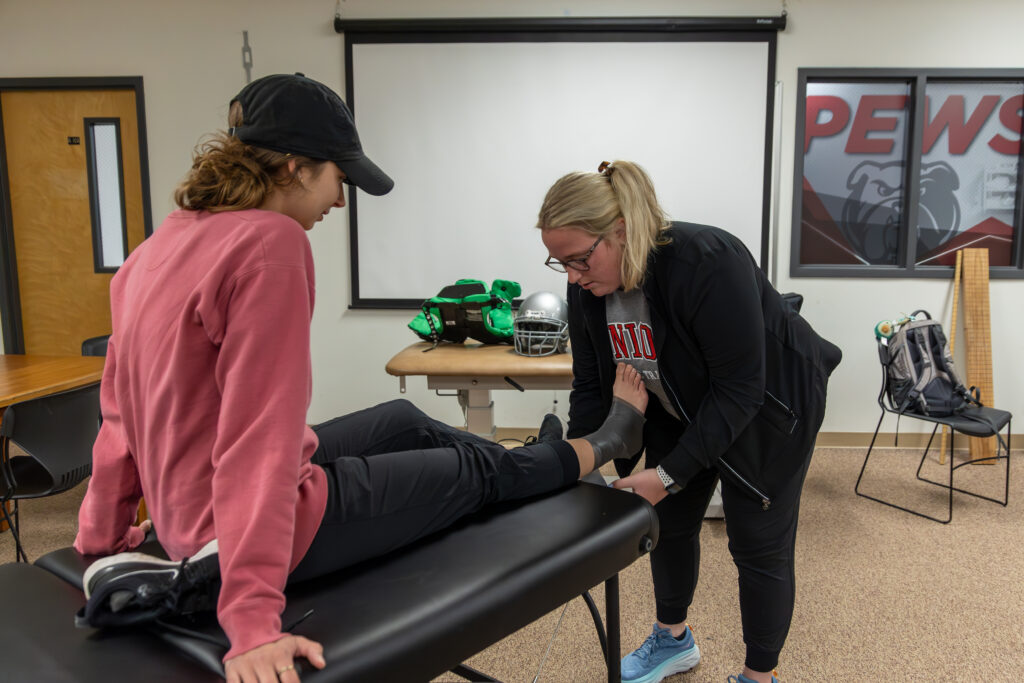 A woman in a pink sweatshirt and black cap sits on an exam table while another woman in glasses examines her ankle in a Pre-Professional Physical Therapy sports training room with equipment visible in the background.