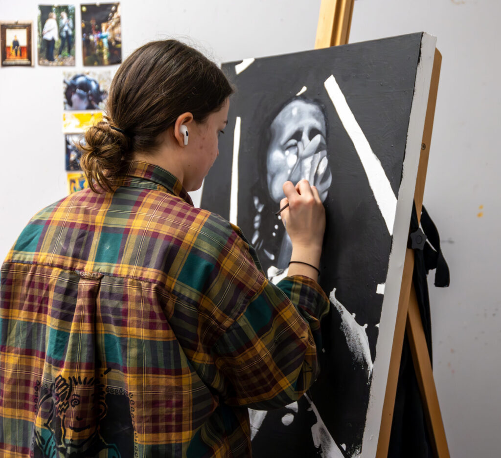 Wearing a plaid shirt and earbud, a person creates an expressive black and white painting on a canvas set on an easel in an art studio. Several photos and images are pinned on the wall, inspiring their art.