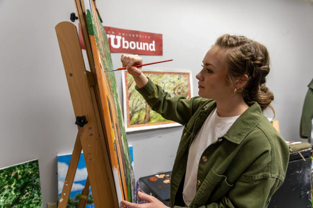 A person wearing a green jacket paints on a canvas at an easel in an art studio. Framed artwork hangs on the wall behind them, and a red sign with UBound is visible.