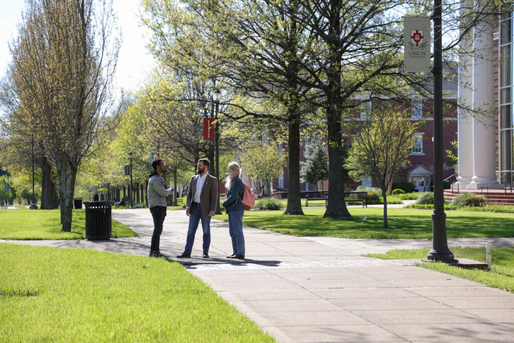 Three people stand talking on a sunny, tree-lined sidewalk at Union University. A banner hangs from a lamp post, promoting the MA in Bible and Theology, while a red brick building completes the vibrant campus scene.