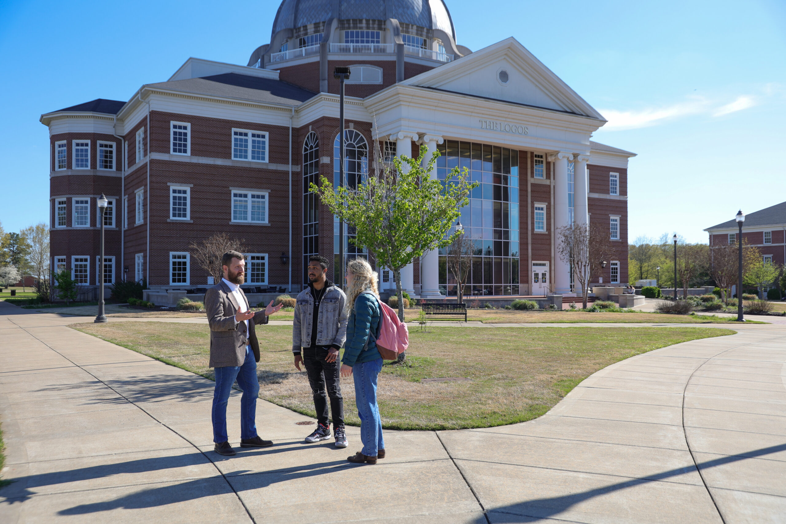 Three people stand talking on a sidewalk in front of a large brick academic building—possibly home to Theology or Religious Studies programs—with tall columns, large windows, and a dome. The sky is clear and the area is landscaped with grass and trees.
