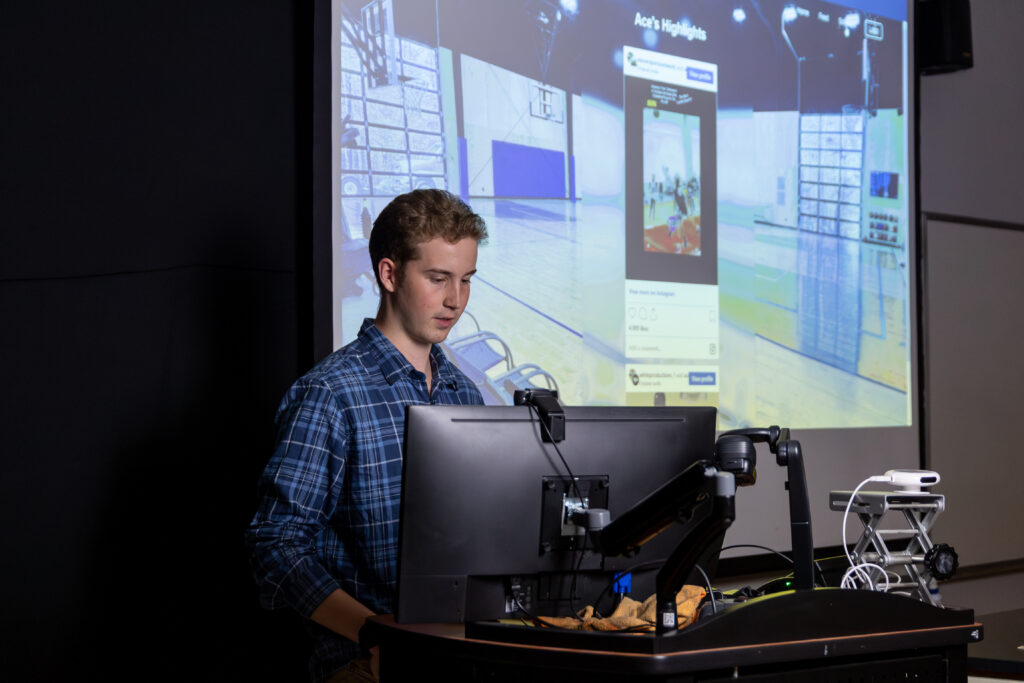 A man in a blue plaid shirt stands at a podium with a computer, giving a presentation on computer science. A large screen behind him displays a gymnasium scene with digital overlays and a highlighted smartphone image.