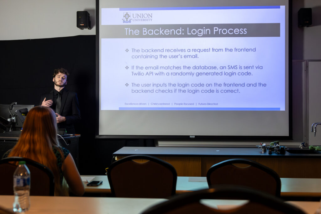 A man stands at the front of a classroom giving a presentation about the backend login process in web development, with a projected slide behind him and students seated at desks facing the presenter.