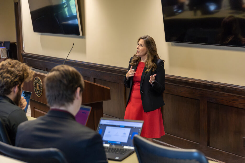 A woman in a red dress and black blazer stands and speaks at the front of a Healthcare Management MBA classroom. Two people with laptops sit facing her, and large monitors are mounted on the wall behind her.