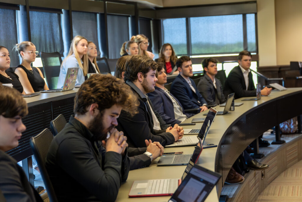 A group of students in business attire sit at curved desks in a classroom, listening attentively to an Operations Management lecture. Most have laptops open, and large windows along the back wall fill the room with natural light.