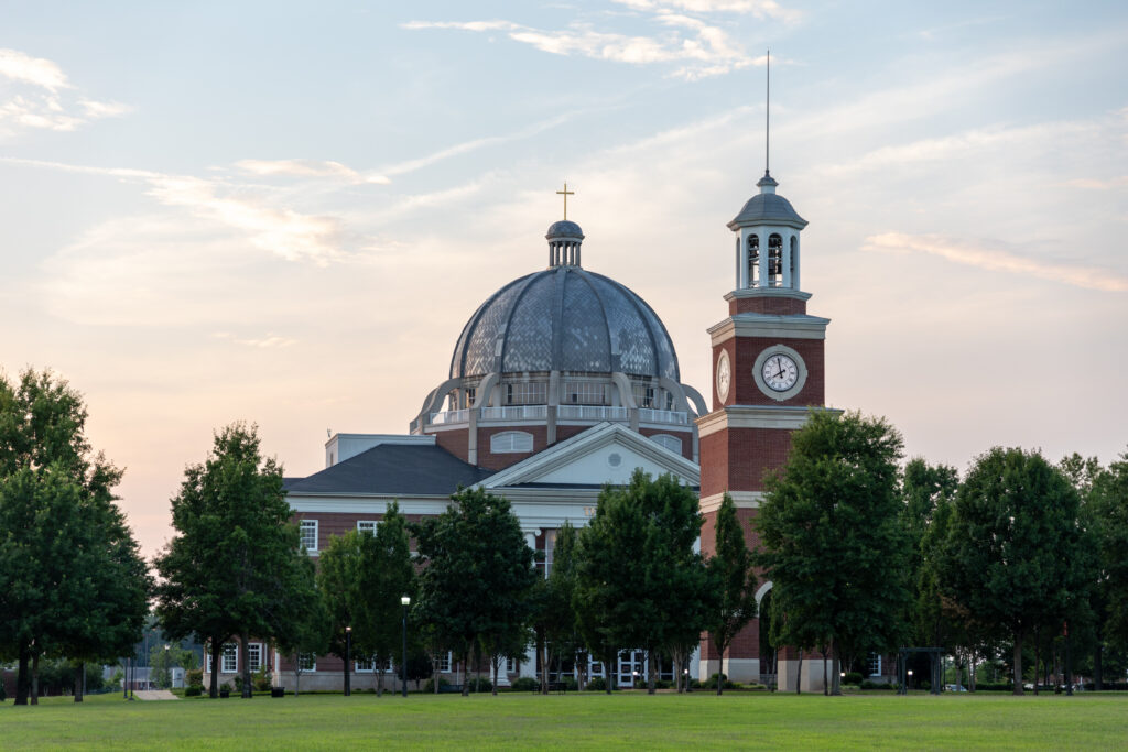 A large brick building with a prominent dome, a clock tower, and a cross on top stands on Union's main campus in Jackson, Tennessee, surrounded by green trees and grass under a partly cloudy sky at sunset.