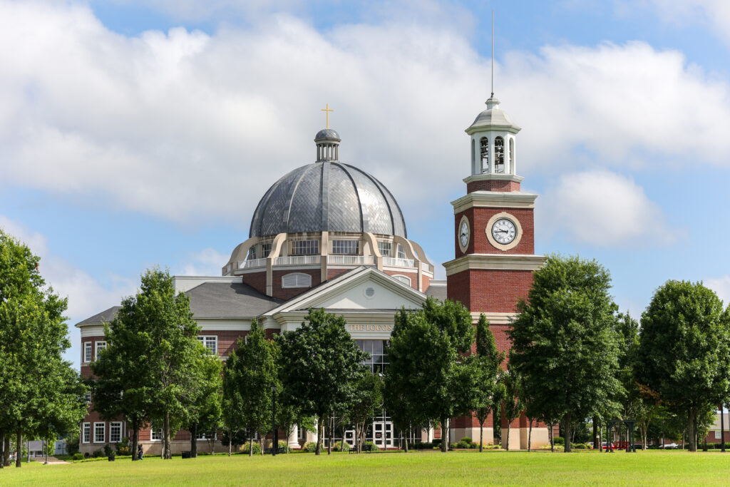 A large brick building with a domed roof and a clock tower, surrounded by green trees and grass, under a partly cloudy sky.