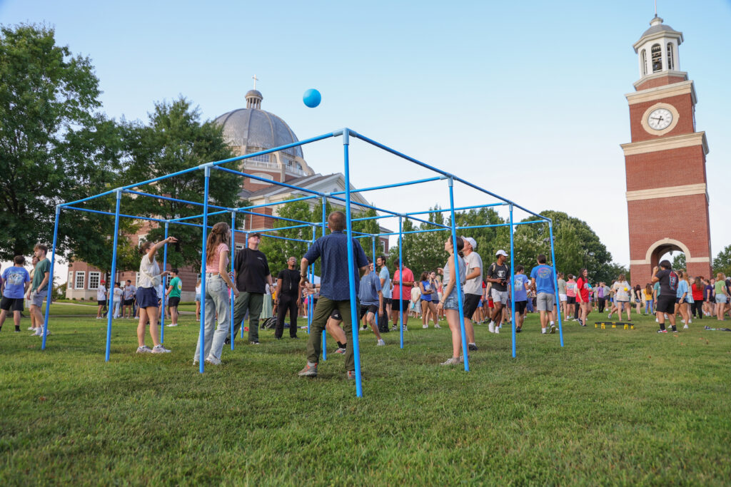 A group of people play a ball game in a blue square structure on a grassy field at Union’s main campus in Jackson, with a domed building and clock tower in the background. Others watch and gather nearby on this sunny Tennessee day.