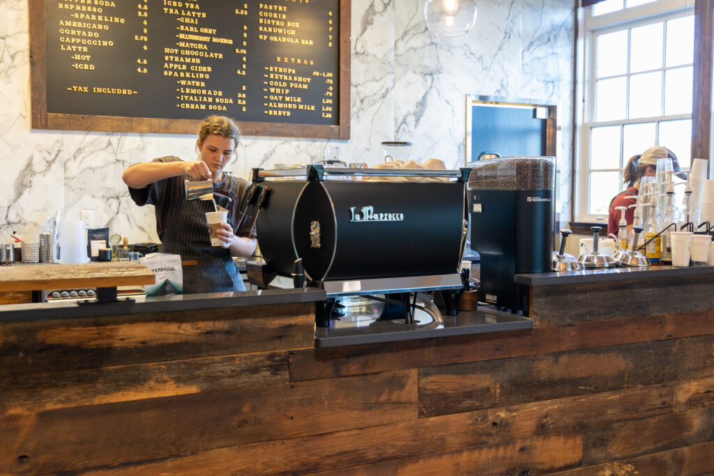 A barista behind a wooden coffee counter pours milk into a cup next to a large espresso machine at Union's main campus in Jackson, Tennessee, with a menu board and stacks of cups visible in the background.