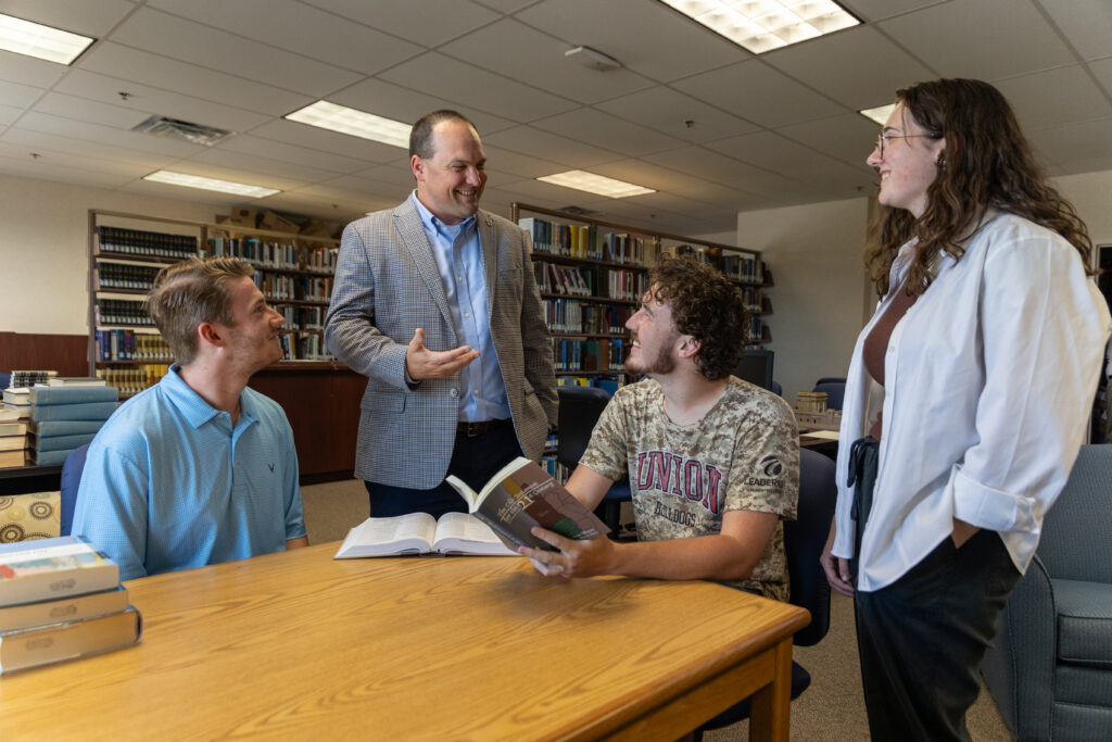 Four people are gathered around a table in a library, engaged in conversation about their Master of Christian Studies. Two are seated with open books, while two stand nearby. Bookshelves filled with books are visible in the background.
