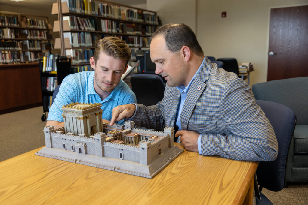 Two men sit at a table in a library, examining a detailed model of an ancient temple—a fitting scene for discussions on intercultural studies or Christian ministry. Bookshelves and a closed wooden door add depth to the scholarly setting.