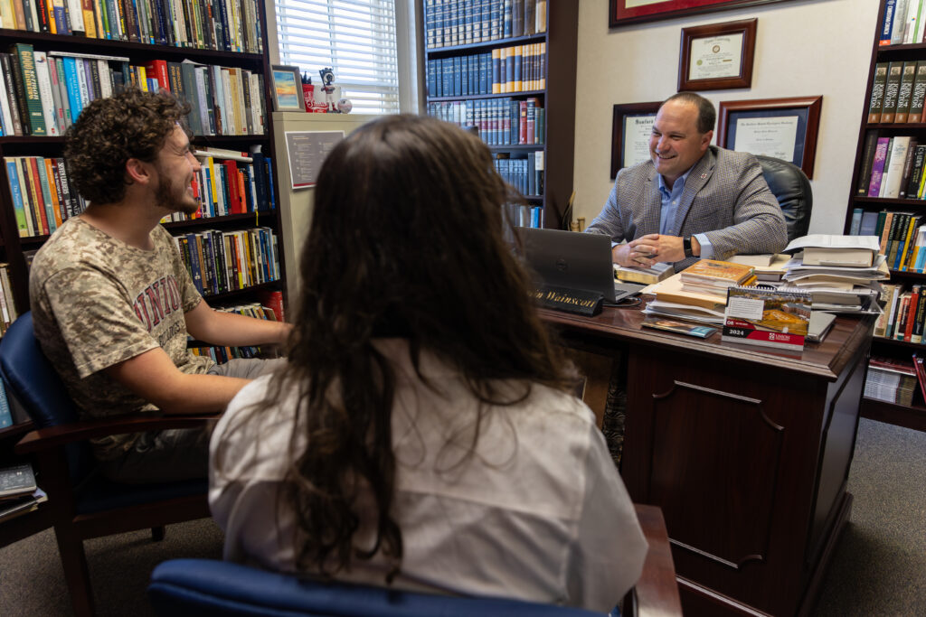 Two students sit across from a man in a suit behind a desk in a book-filled office. The man, engaged in Biblical Counseling, is smiling and speaking with the students, who appear to be listening attentively.