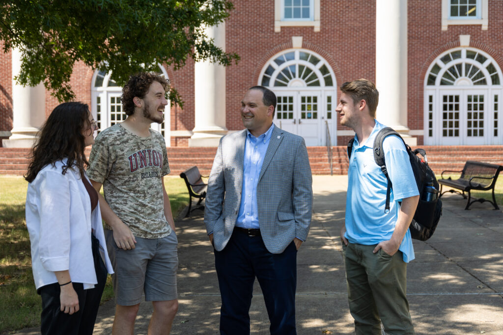 Four people stand outside a brick building with arched windows, talking. Three appear to be students, one is an adult in a blazer. They are on a paved path with benches and trees visible.