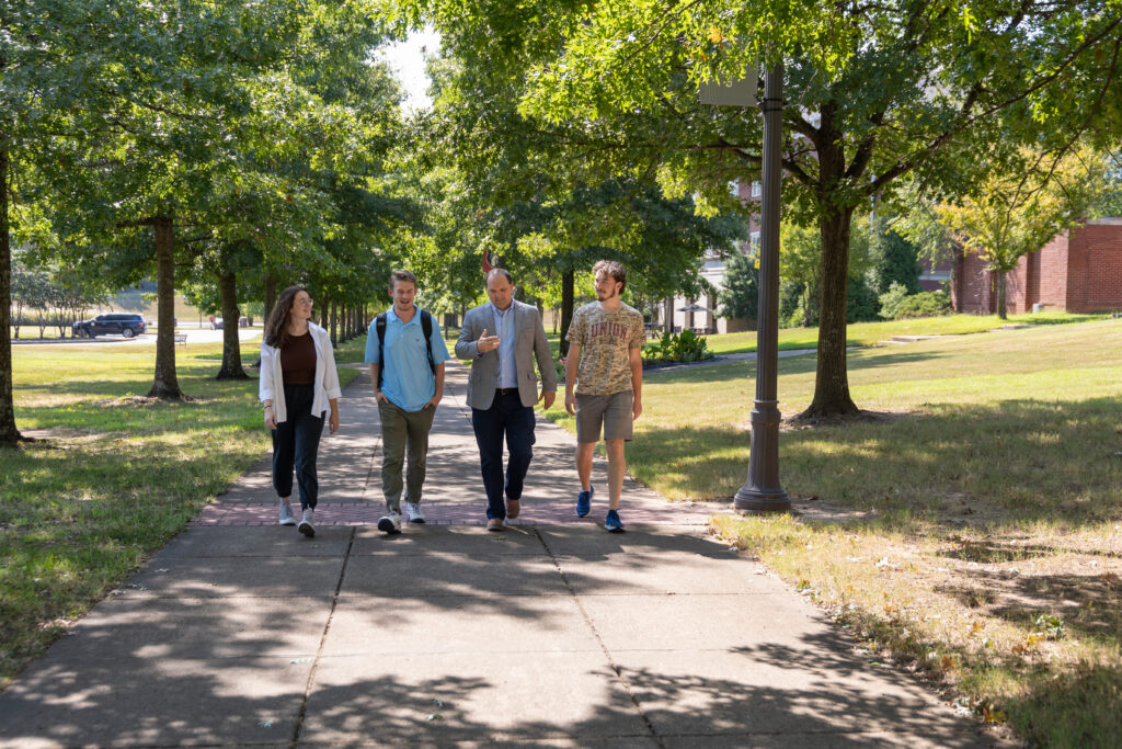 Four people walk together down a sunny, tree-lined sidewalk. One in business attire gestures while talking about Youth Ministry to the others, who are casually dressed with backpacks, creating a lively discussion about their shared mission.