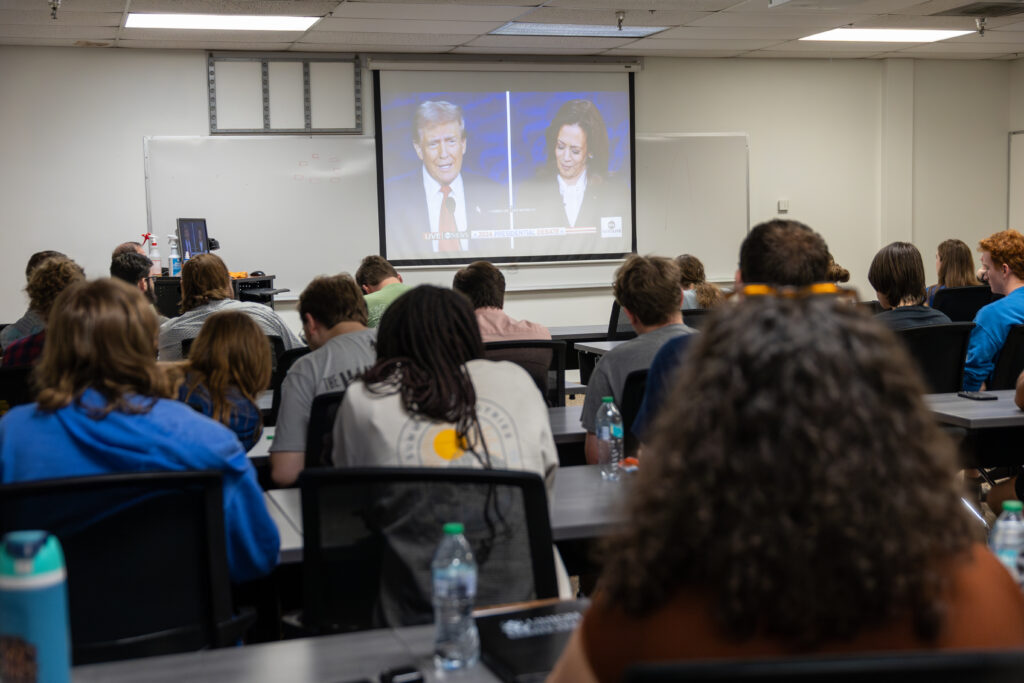 Students sit in a classroom facing a projector screen displaying an image of two people speaking during a televised broadcast on American Politics. Bottles and notebooks are visible on the desks.