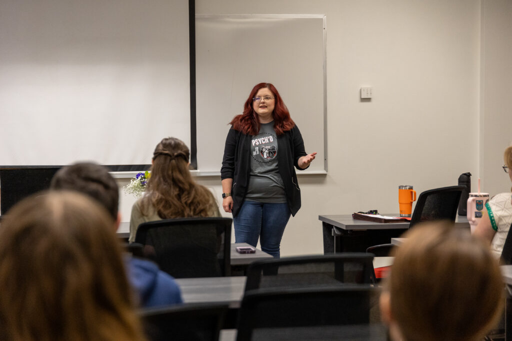 A woman with red hair stands at the front of a classroom, speaking to seated students. She is wearing jeans, a gray t-shirt, and a black jacket. A whiteboard and a projector screen are visible behind her.