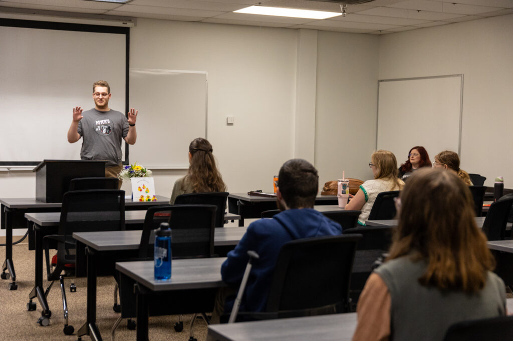 A man stands at the front of a classroom speaking to several seated students. The students face him, and desks are arranged in rows. The room is well-lit, with white walls and a whiteboard behind the speaker.
