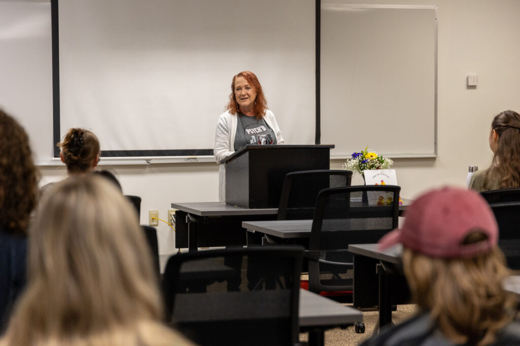 A woman stands at a podium speaking to a small audience in a classroom. There are desks, chairs, a flower arrangement, and a whiteboard in the background. The audience listens attentively.