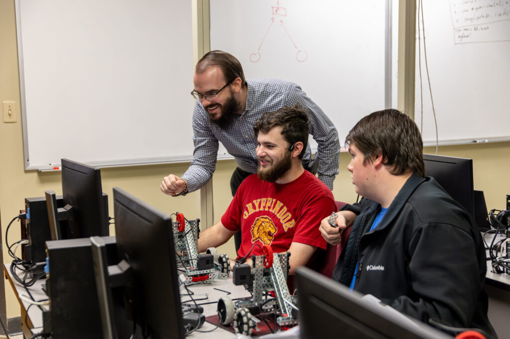 A bearded man stands and smiles while observing two seated men working with robotics equipment and computers in a classroom, exploring concepts in Computer Science and Machine Learning. Whiteboards with diagrams are visible in the background.
