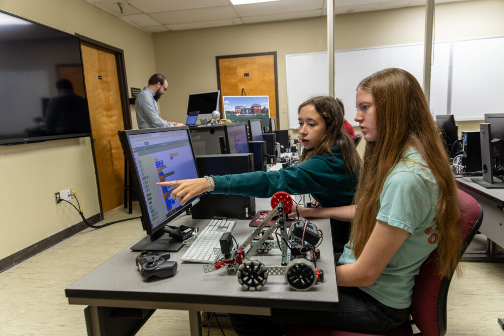 Two young women sit at a desk using a computer, with robotics equipment and a controller in front of them—one points at the monitor, possibly analyzing data science results. Another person stands in the background, working at a separate desk.