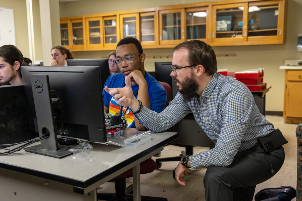 A man crouches beside a seated student, pointing at a computer screen in a classroom where Computer Science is taught. Other students work at computers in the background amid wooden cabinets and various supplies.