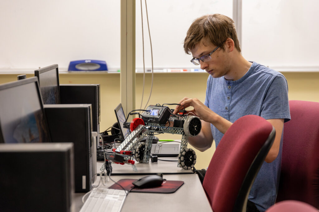 A person in glasses and a blue shirt sits at a desk assembling a metal robot with wheels, surrounded by computer monitors and office chairs in a Computer Science classroom or lab setting.