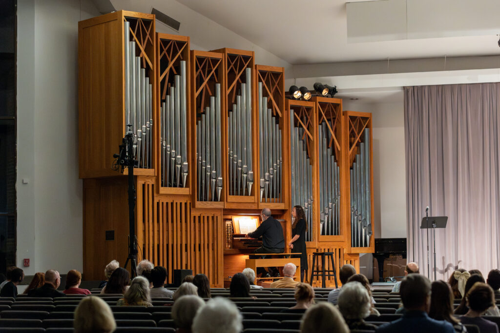 A musician delivers a captivating music performance on a grand pipe organ in front of an audience in a church or concert hall. The organ’s tall metal pipes and wooden structure stand out as people watch the performer.