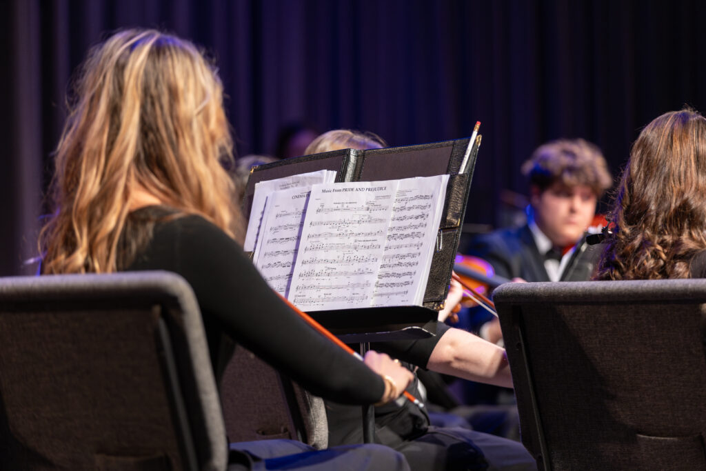 A person with long hair is seated in front of a music stand with sheet music, playing a string instrument in an orchestra. The scene reflects the art of composition, with other musicians and chairs visible in the background.