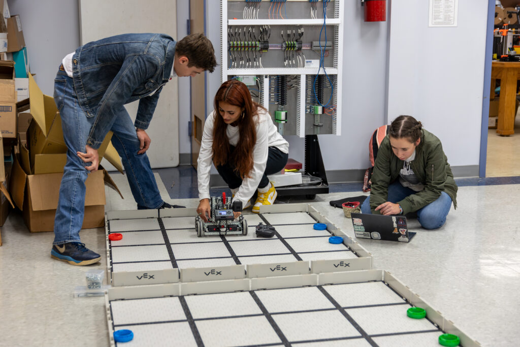 Three students collaborate on an engineering project; one adjusts a small robot on a VEX competition field, another observes closely, and the third—an aspiring electrical engineer—works on a laptop amid equipment in the background.
