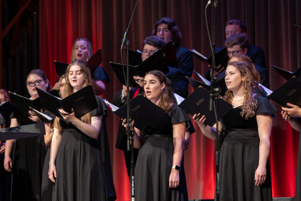 A group of choir singers, dressed in black formal attire, stand in rows holding open music folders and deliver a captivating music performance on stage with microphones, set against red and brown illuminated curtains.
