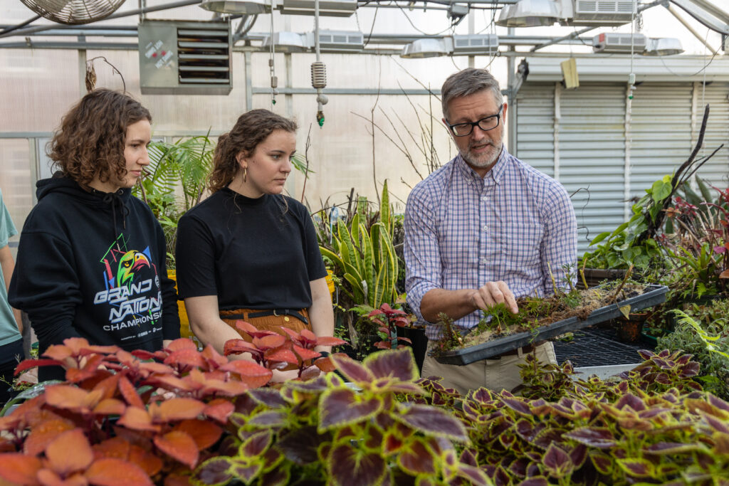 A man shows two women a tray of small plants inside a greenhouse filled with various leafy plants. The women watch attentively as the man explains something about the tray’s contents.