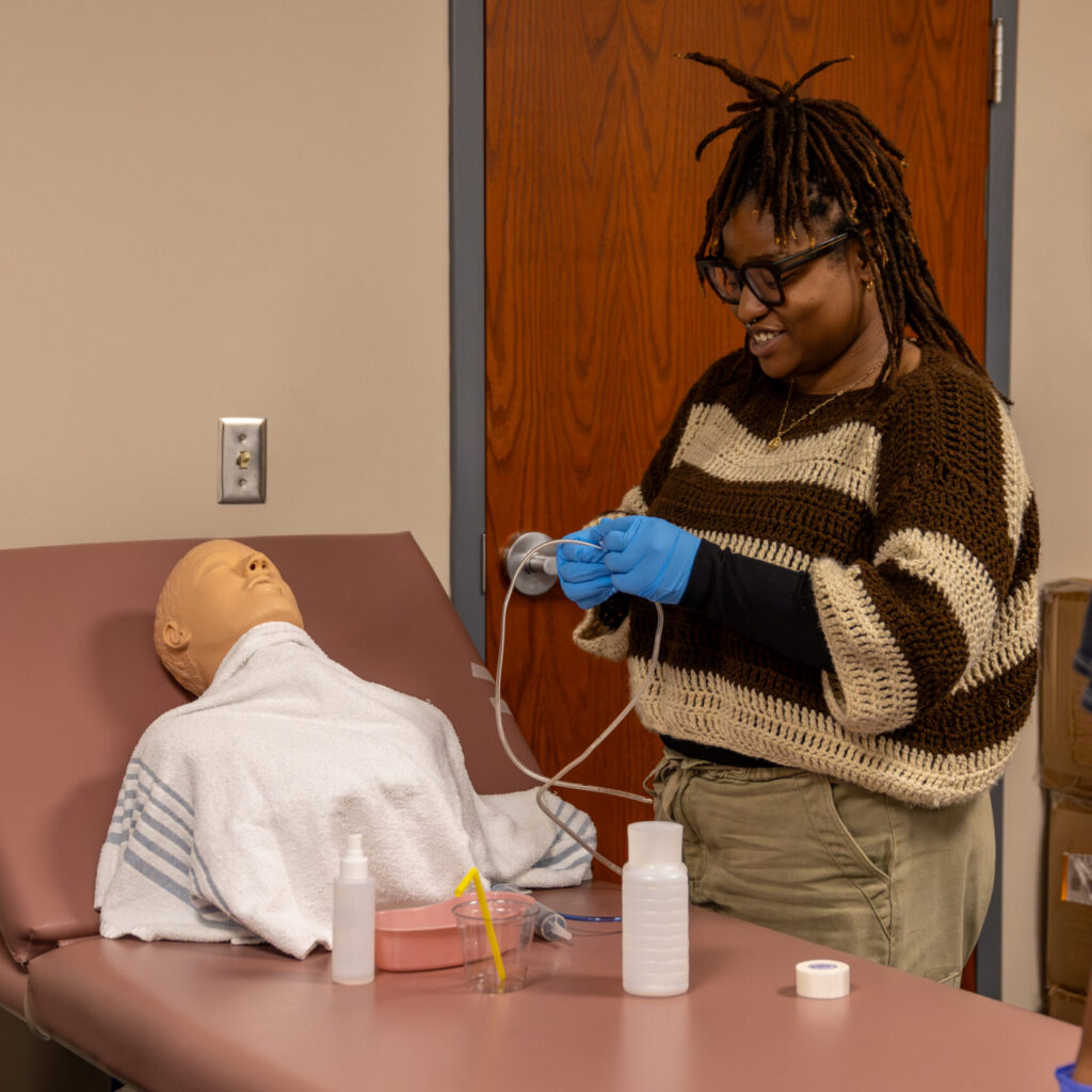 A person wearing gloves prepares medical equipment next to a medical mannequin lying on an examination table, simulating a clinical procedure in a training room at the Memphis Campus.