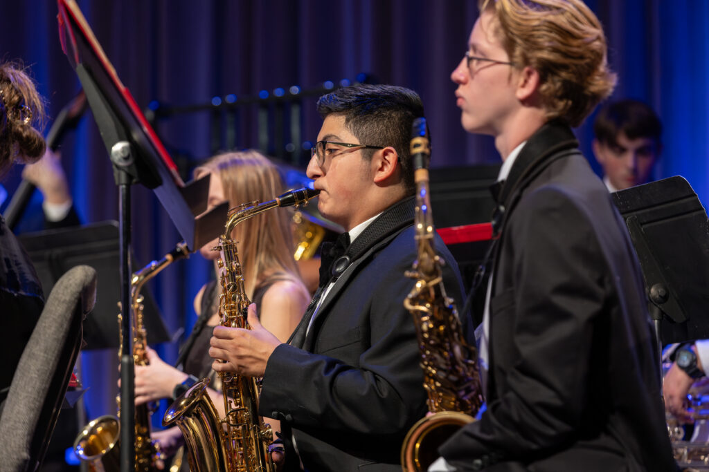 A group of musicians in formal black attire give an instrumental performance, playing saxophones while seated in rows with music stands before them on a stage set against a purple curtain backdrop, showcasing the importance of music education.